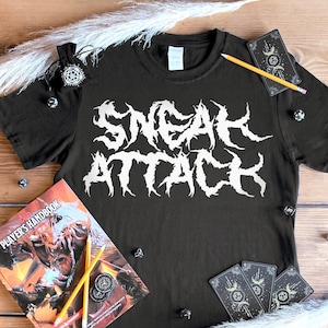 Puede incluir: Camiseta negra con el texto blanco "SNEAK ATTACK". La camiseta está rodeada por un manual del jugador, dados, cartas del tarot, un lápiz y elementos decorativos. Los artículos están dispuestos sobre una superficie de madera.