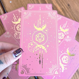 Puede incluir: Cartas de tarot rosas con diseños celestiales dorados, que incluyen lunas, estrellas y formas geométricas. Las cartas están dispuestas sobre una superficie de madera, mostrando la obra de arte detallada y el tema místico.