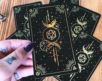 May include: Black tarot cards with gold foil celestial designs. The cards feature moons, stars, and geometric shapes. The cards are held in a hand with glitter nail polish. The cards are on a wooden surface.