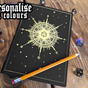 May include: A black notebook with a gold geometric design and the text "personalise foil colours". An orange pencil rests on the notebook. Dice and star-shaped confetti are scattered on the wooden surface.