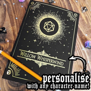May include: Black journal with gold foil accents, featuring a D20 die symbol and the text "Willow Whisperwind." Includes an orange pencil and dice scattered around. The journal is designed to be personalized with any character name.
