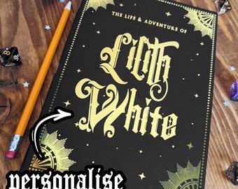 Puede incluir: Un diario negro con letras doradas que dice "The Life & Adventure of Lilth White". El diario tiene un borde decorativo y detalles de estrellas. Un lápiz y dados están al lado del diario. El texto "personalise with any character name!" también es visible.