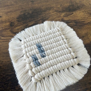 May include: A square woven coaster with a fringe border. The coaster is made of white cotton rope and has a blue and white geometric design in the center.