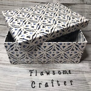 Puede incluir: Una caja blanca con un patrón geométrico azul y blanco. La tapa está abierta y la caja está sobre una superficie de madera blanca. El texto "Flawesome Crafter" es visible en la superficie.