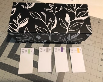 May include: Four small white cards with colour swatches and DMC thread numbers. The cards are labelled with the DMC thread numbers 100, 109, 121, and 130. The colour swatches are green, grey, purple, and yellow. The cards are sitting on a black and white patterned box.