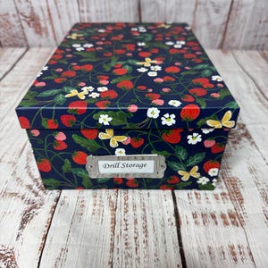 May include: A rectangular storage box with a navy blue background, adorned with a pattern of red strawberries, white flowers, and yellow butterflies. The box has a metal label that reads "Drill Storage".