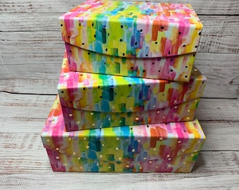 May include: Four colourful boxes stacked on top of each other. The boxes are decorated with a watercolour-style pattern of pink, yellow, blue, and green brushstrokes. The boxes have a white background and gold polka dots.