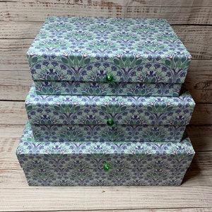 May include: Three decorative boxes stacked on top of each other. The boxes are covered in a floral pattern with shades of blue, green, and purple. Each box has a green knob.