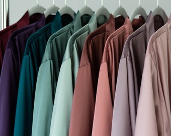 May include: A row of silk robes in various colors, including burgundy, purple, teal, sage green, and mauve, hang on a clothing rack. The robes have a satin finish and are displayed against a neutral background. The robes are open-fronted with long sleeves.