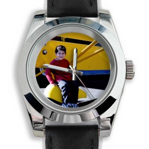 May include: A silver wristwatch with a black leather band. The watch face features a photograph of a child in a red shirt sitting on a yellow airplane. The watch has silver hands and a silver bezel.