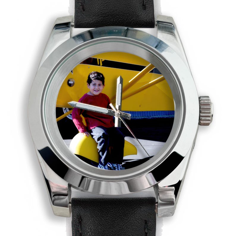 May include: A silver wristwatch with a black leather band. The watch face features a photograph of a child in a red shirt sitting on a yellow airplane. The watch has silver hands and a silver bezel.