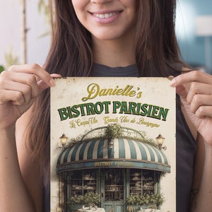 Może przedstawiać: W stylu vintage znak dla francuskiego bistro o nazwie "Danielle's Bistrot Parisien." Znak zawiera zielony daszek w białe pasy i przedstawia paryski sklep z stolikami i krzesłami na zewnątrz. Tekst "Le Coq au Vin Grands Vins de Bourgogne" jest napisany ozdobną czcionką pod nazwą bistro. Znak zawiera r&oacute;wnież tekst "Glace bar a vin" na dole.