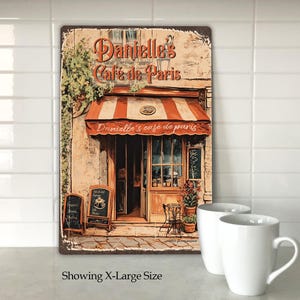 Puede incluir: Un letrero de metal de estilo vintage que representa un caf&eacute; parisino con una marquesina roja y el texto "Danielle's Cafe de Paris". El letrero tiene un aspecto desgastado y presenta un edificio de ladrillo con una puerta y ventanas.