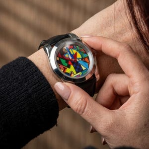May include: A stainless steel wristwatch with a black leather band. The watch face features a colorful abstract design with red, yellow, blue, and green shapes. The watch is being worn on a wrist.