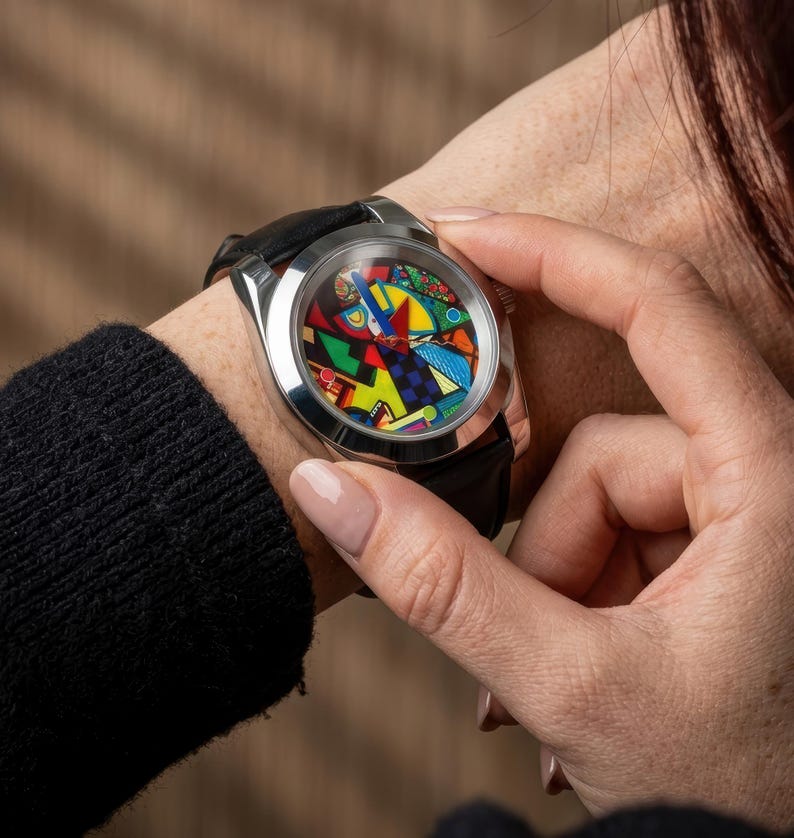 May include: A stainless steel wristwatch with a black leather band. The watch face features a colorful abstract design with red, yellow, blue, and green shapes. The watch is being worn on a wrist.