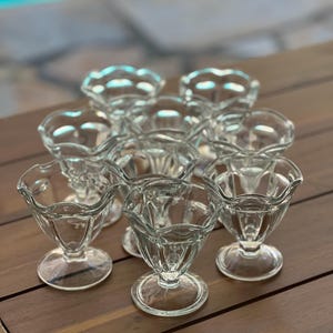 May include: A collection of clear glass dessert dishes with scalloped edges and pedestal bases. The dishes are arranged on a dark wooden surface, with a blurred background of a pool and stone patio.
