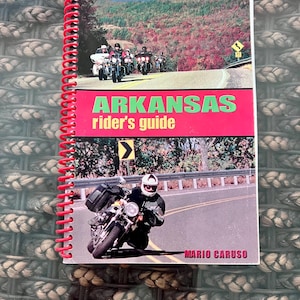 Puede incluir: Un libro encuadernado en espiral titulado "ARKANSAS rider's guide". La portada muestra motocicletas en una carretera sinuosa, con el título en verde y rojo. El nombre del autor, Mario Caruso, es visible en la parte inferior.