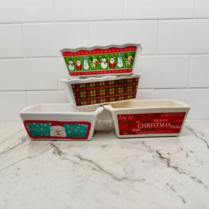 May include: Four rectangular ceramic loaf pans with festive designs. One features snowmen and Santa, one has a red and green plaid pattern, one shows a Santa face with candy canes, and one displays red text reading Joy to the World Christmas.