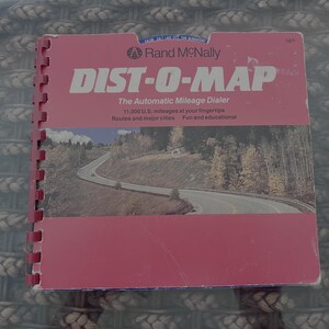 May include: A vintage Rand McNally DIST-O-MAP, a spiral-bound book with a red cover. The cover features the title in large white letters and a scenic road illustration. Text includes "The Automatic Mileage Dialer" and "11,000 U.S. mileages at your fingertips."