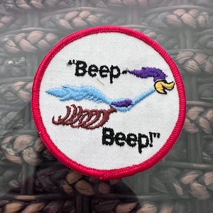 May include: Circular patch with a white background and a red border. The patch features a cartoon roadrunner in blue, purple, and yellow, with the words "Beep Beep!" in black.