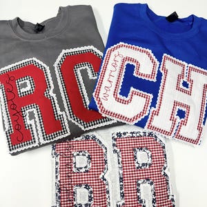 Pode incluir: Três camisolas com grandes apliques de letras. Uma camisola cinzenta tem um "R" vermelho e a palavra "coyotes". Uma camisola azul tem um "C" branco e a palavra "warriors". A terceira camisola tem um aplique "B" e "R" xadrez vermelho e branco.