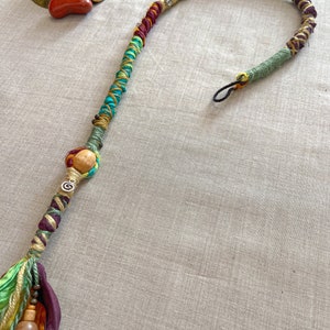May include: A colourful braided hair extension with wooden beads and a metal charm. The extension is made of various colours of yarn and fabric, including green, yellow, orange, red, and brown.