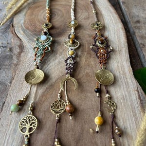 May include: Three macrame hair accessories with brass accents and decorative charms. Each features a unique design with beads, spiral discs, and symbols like a tree of life, lotus flower, and Om symbol. The colour palette includes brown, gold, and earthy tones.
