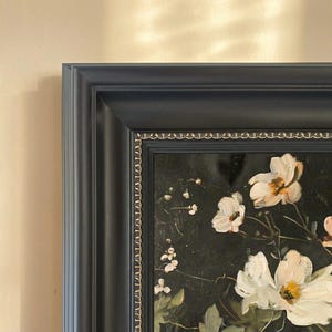 Pode incluir: Pintura floral emoldurada com flores brancas e centros amarelos sobre um fundo escuro. A pintura está emoldurada em uma moldura azul escura com uma borda interna decorativa. A obra de arte é exibida em uma parede bege.