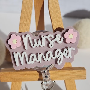 Peut inclure: Un porte-badge avec les mots "Nurse Manager" en écriture blanche sur un fond lavande. Le badge est décoré d'accents de fleurs roses et d'un enrouleur rétractable en plastique transparent avec un clip en métal.