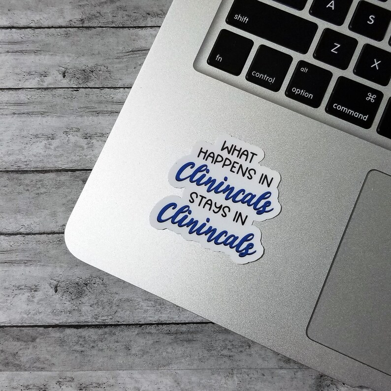 May include: A white sticker with the text "WHAT HAPPENS IN Clinicals STAYS IN Clinicals" in blue script lettering. The sticker is on a silver laptop, with a wooden background.