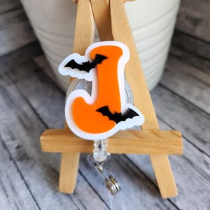May include: Orange letter J with black bats on a white background. The letter is attached to a retractable badge reel.