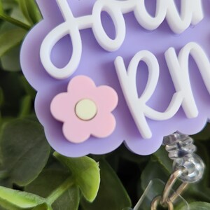 May include: A light purple badge reel with the name "Joan RN" in white script. A pink flower with a yellow center is on the bottom left. The badge reel has a clear plastic clip and a silver metal retractable cord.