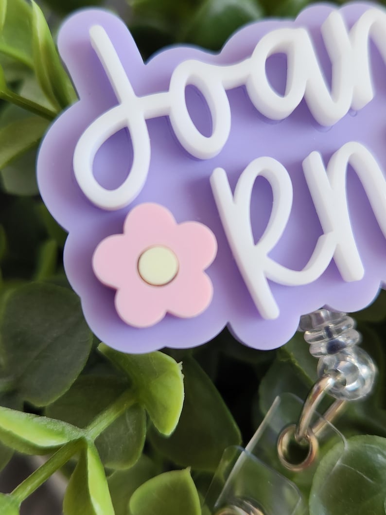 May include: A light purple badge reel with the name "Joan RN" in white script. A pink flower with a yellow center is on the bottom left. The badge reel has a clear plastic clip and a silver metal retractable cord.