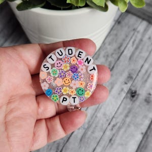 Puede incluir: Un portagafete redondo de color rosa y blanco con las palabras "STUDENT PT" escritas en cuentas blancas. El portagafete est&aacute; decorado con flores de colores y caritas sonrientes.