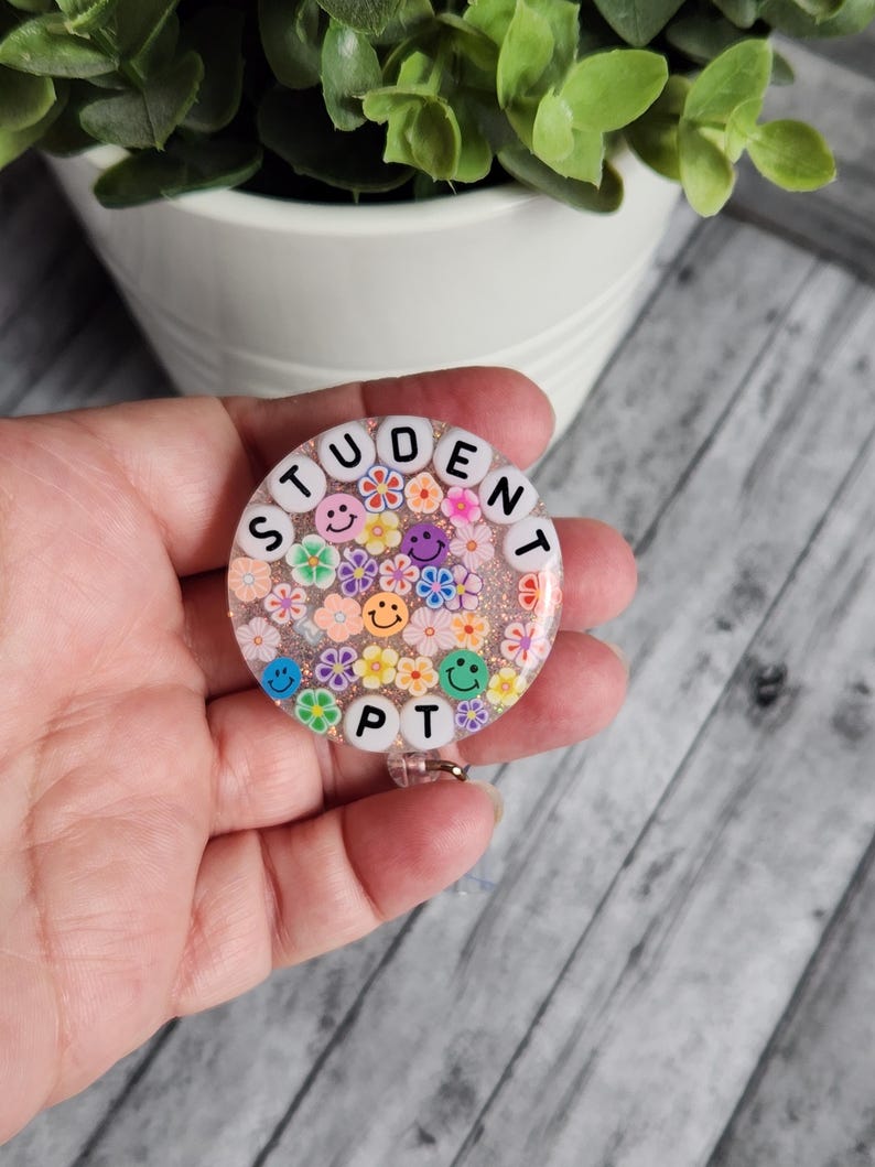 Puede incluir: Un portagafete redondo de color rosa y blanco con las palabras "STUDENT PT" escritas en cuentas blancas. El portagafete est&aacute; decorado con flores de colores y caritas sonrientes.