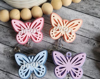 May include: Four butterfly-shaped badge reels in pink, peach, blue, and purple. Each butterfly has the name "Joan" in white script. The badge reels are on a clear plastic base with a metal clip.