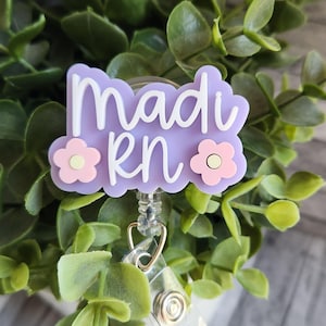 May include: A light purple badge reel with the name "Madi RN" in white script. The badge has a floral design with pink and white flowers. The reel is attached to a clear plastic holder and a metal clip.