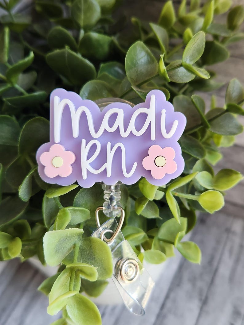 May include: A light purple badge reel with the name "Madi RN" in white script. The badge has a floral design with pink and white flowers. The reel is attached to a clear plastic holder and a metal clip.