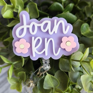 May include: A light purple badge reel with the name "Joan RN" in white script. The badge has two pink flower accents and a clear plastic retractable clip. The background is a bed of green leaves.