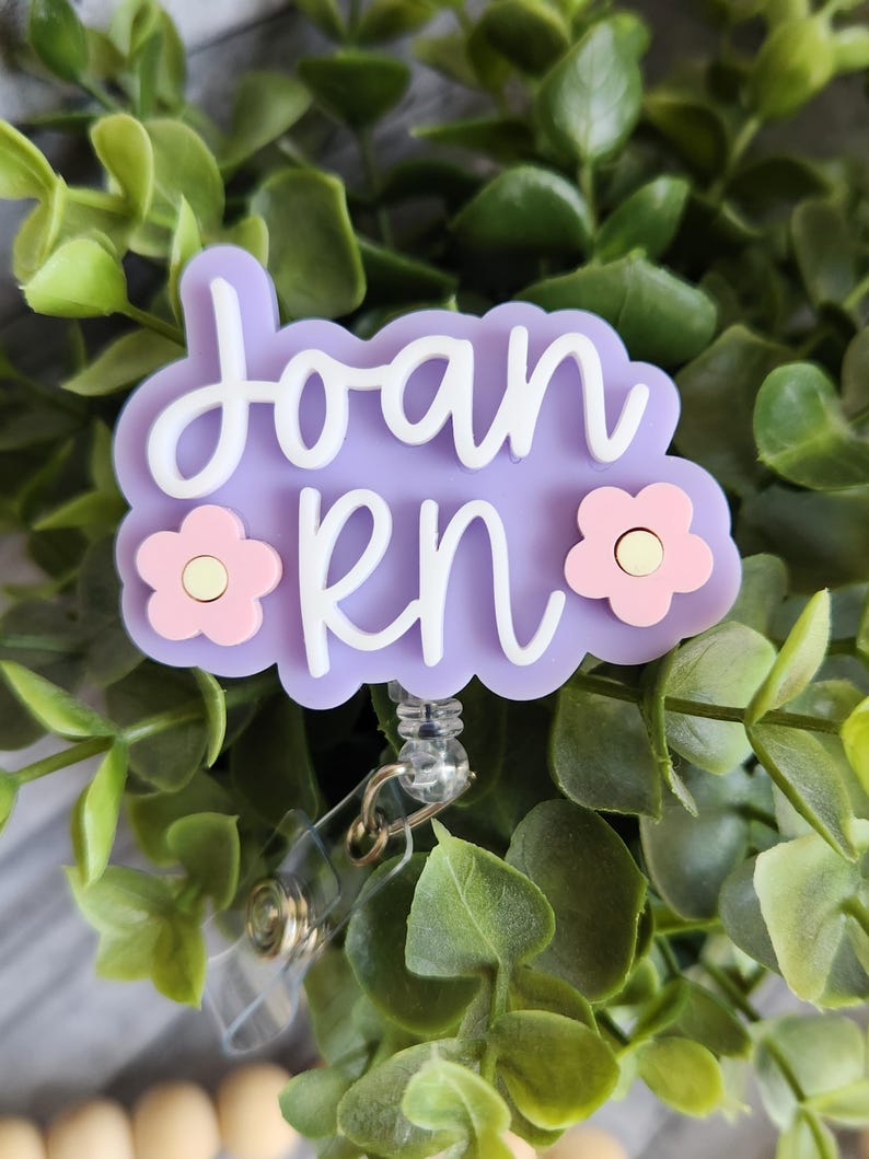 May include: A light purple badge reel with the name "Joan RN" in white script. The badge has two pink flower accents and a clear plastic retractable clip. The background is a bed of green leaves.