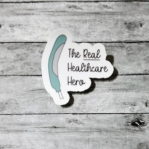 May include: A white sticker with a teal and gray medical device illustration and the text "The Real Healthcare Hero" in black. The sticker is on a gray and white wooden surface.