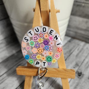 Puede incluir: Un portagafete blanco y con brillantina con un inserto de gafete de pl&aacute;stico transparente. El gafete tiene un fondo blanco con flores de colores y caras sonrientes. El texto "STUDENT PT" est&aacute; escrito en letras blancas alrededor del borde del gafete.