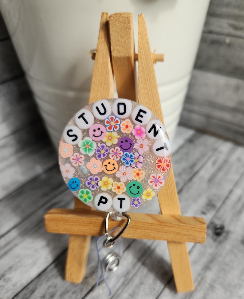Puede incluir: Un portagafete blanco y con brillantina con un inserto de gafete de pl&aacute;stico transparente. El gafete tiene un fondo blanco con flores de colores y caras sonrientes. El texto "STUDENT PT" est&aacute; escrito en letras blancas alrededor del borde del gafete.