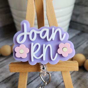 May include: A light purple RN badge reel with the name "Joan" in white script. The badge has a cloud shape with two pink flower accents. The reel is attached to a silver clip.
