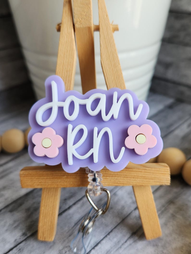May include: A light purple RN badge reel with the name "Joan" in white script. The badge has a cloud shape with two pink flower accents. The reel is attached to a silver clip.
