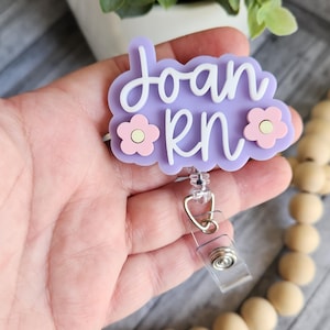May include: A light purple badge reel with the name "Joan RN" in white script. The badge has a floral design with pink flowers and a clear plastic retractable reel clip.