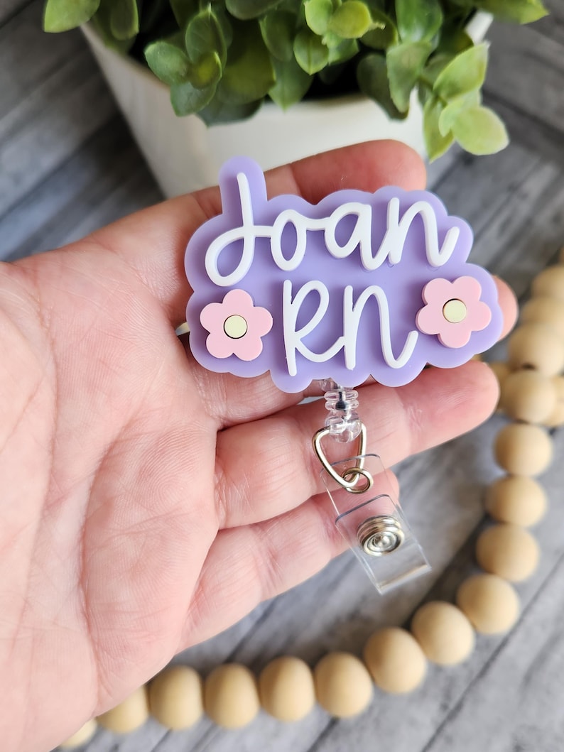 May include: A light purple badge reel with the name "Joan RN" in white script. The badge has a floral design with pink flowers and a clear plastic retractable reel clip.