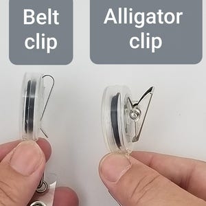 May include: Two clear plastic badge reels are shown, one with a belt clip and the other with an alligator clip. The text "Belt clip" and "Alligator clip" are displayed above each reel. The reels are held by hands.