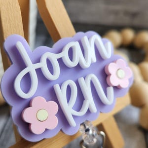 May include: A light purple badge reel with the name "Joan RN" in white script. The badge has a cloud-like shape and is decorated with two pink flower accents. The reel is attached to a clear plastic clip.