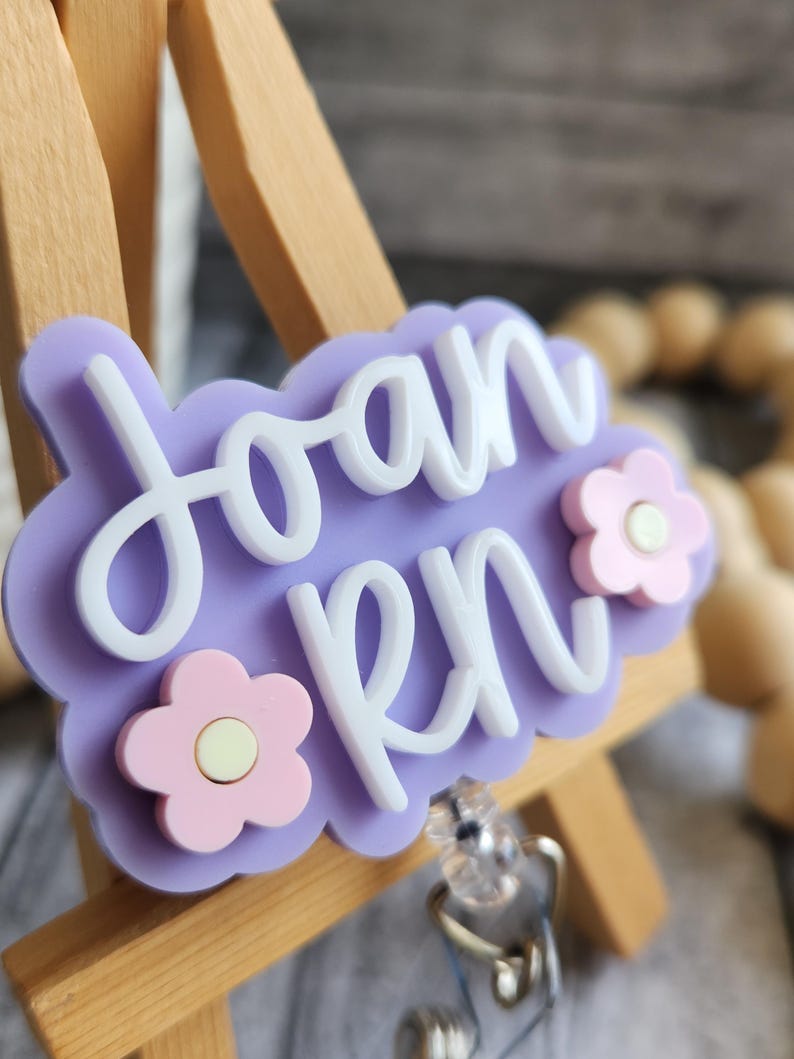May include: A light purple badge reel with the name "Joan RN" in white script. The badge has a cloud-like shape and is decorated with two pink flower accents. The reel is attached to a clear plastic clip.